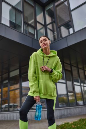 low angle view, sportswoman in oversized hoodie and leggings standing with bottle with water outdoor