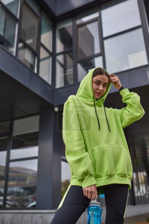young sportswoman in oversized active wear wearing hood and standing with bottle with water outdoor