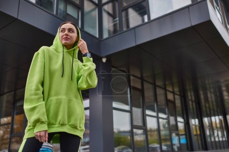junge Sportlerin in Kapuzenpulli und Leggings steht mit Flasche mit Wasser in der Nähe moderner Gebäude