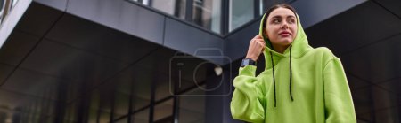 Banner einer jungen Sportlerin mit Kapuze auf dem Kopf, die in der Nähe eines modernen Gebäudes steht, lindfarbener Kapuzenpullover
