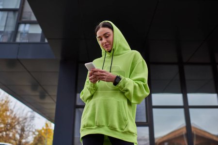 junge brünette Sportlerin mit Kapuze auf dem Kopf mit Smartphone in der Nähe moderner Gebäude, Straßenstil