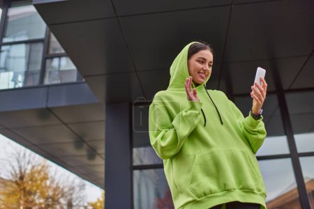 happy young woman in hoodie waving hand while having video call on smartphone near modern building