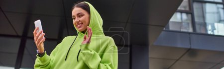 mujer joven feliz en sudadera con capucha saludando la mano mientras tiene videollamada en el teléfono inteligente cerca del edificio, pancarta