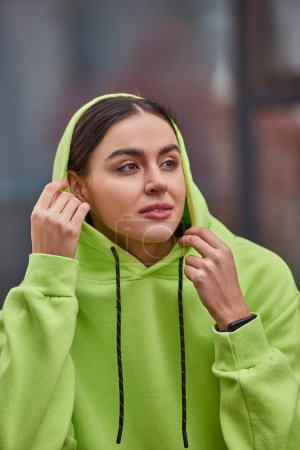 brünette junge Frau in lindfarbenem Kapuzenpulli, Kapuze auf dem Kopf und Blick nach draußen