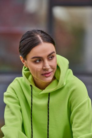 portrait of dreamy young woman with brunette hair in lime color hoodie looking away outdoors
