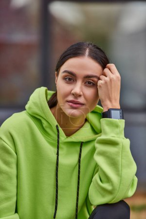 pretty young woman in lime color hoodie posing with smart watch on wrist and looking at camera