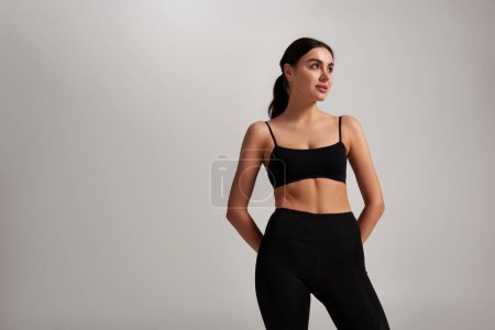 beautiful and young sportswoman in black leggings and crop top standing on grey background