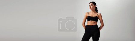 beautiful and young sportswoman in black leggings and crop top standing on grey background, banner