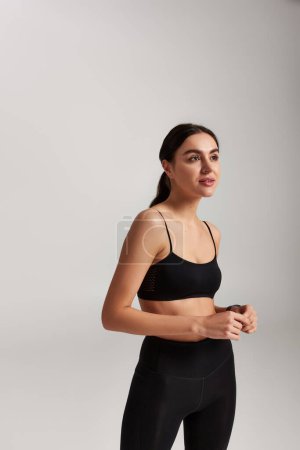 verträumte junge Sportlerin in schwarzen Leggings und bauchfreiem Top auf grauem Hintergrund
