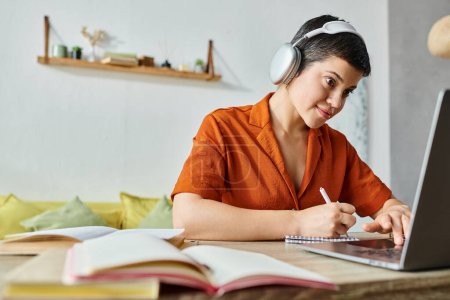 fröhliche hübsche Frau in lässigem Outfit mit Kopfhörern, die Notizen macht und auf Laptop schaut, studiert
