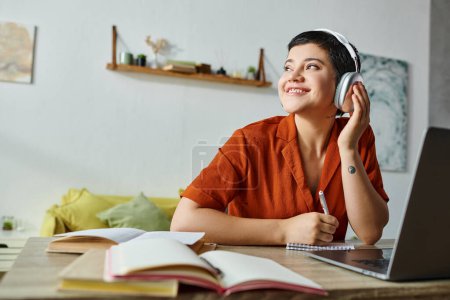 Photo for Cheerful young student in orange shirt listening music while studying at laptop, looking away - Royalty Free Image