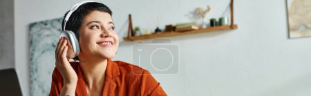 Photo for Jolly student in orange shirt with headphones studying at laptop, looking away, education, banner - Royalty Free Image