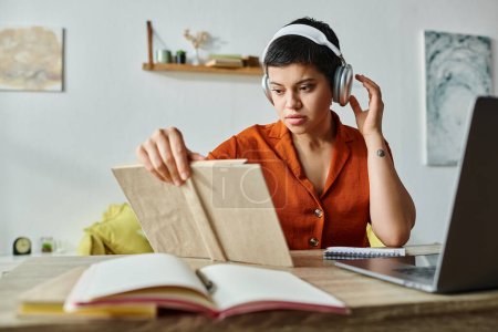 Téléchargez les photos : Jeune femme attrayante avec les cheveux courts et écouteurs livre de lecture tout en étudiant à la maison, éducation - en image libre de droit