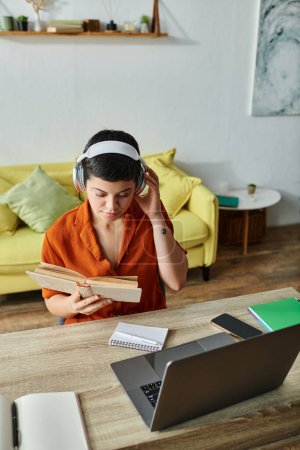 Vertikale Aufnahme einer nachdenklichen Frau in Freizeitkleidung, die von zu Hause aus mit Lehrbuch und Laptop lernt