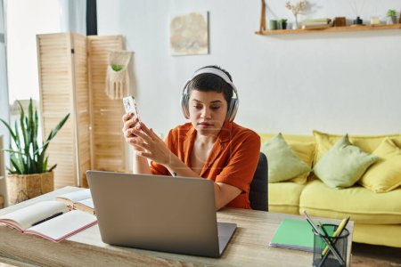 Téléchargez les photos : Jeune femme aux cheveux courts avec écouteurs et téléphone portable étudiant à son ordinateur portable à la maison, éducation - en image libre de droit
