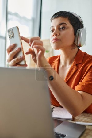 vertikale Aufnahme eines attraktiven, kurzhaarigen Studenten mit Kopfhörern, der vor dem Laptop auf das Telefon schaut