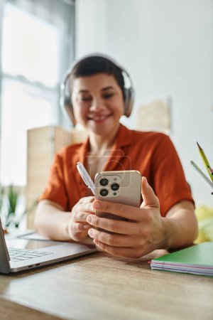 Photo for Vertical focused shot of phone in joyful female student hands, headphones and laptop, education - Royalty Free Image