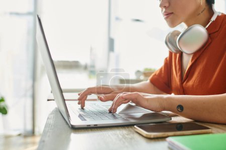 cropped view of young woman in orange shirt with tattoo and headphones studying at laptop, education