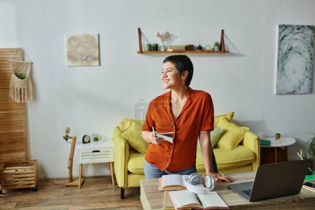 Photo for Happy young student in casual clothes standing next to her desk with laptop and notes, looking away - Royalty Free Image