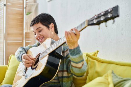 Foto de Alegre atractiva mujer con pelo corto en remoto lección de guitarra sonriendo felizmente, educación en casa - Imagen libre de derechos