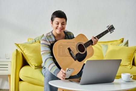 Photo for Cheerful attractive woman looking at her phone during remote guitar lesson, education at home - Royalty Free Image