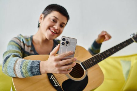 junge attraktive Frau blickt freudig auf Handy mit Gitarre in der Hand, Ausbildung zu Hause