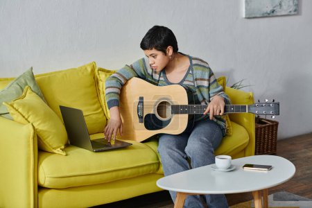 Photo for Attractive young woman sitting on sofa with guitar in hands and looking at laptop, education at home - Royalty Free Image