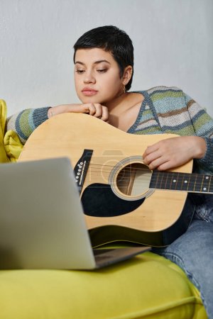 vertikale Aufnahme einer jungen attraktiven Frau, die auf dem Sofa sitzt und an einer Online-Gitarrenstunde teilnimmt