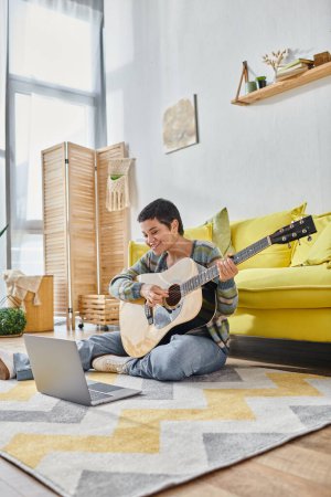 Téléchargez les photos : Plan vertical de la femme assise sur le sol avec la guitare souriant à la caméra de l'ordinateur portable, éducation à la maison - en image libre de droit