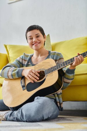 Photo for Vertical shot of cheerful woman sitting on floor with guitar smiling at camera, education at home - Royalty Free Image