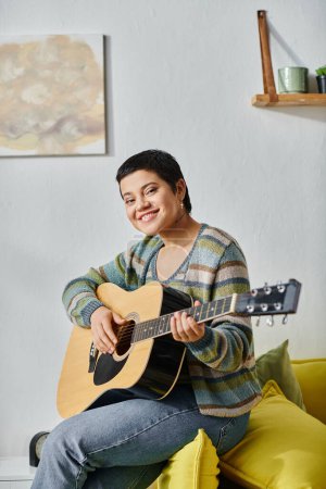 Photo for Vertical shot of smiling happy woman in casual attire playing guitar on sofa, education at home - Royalty Free Image
