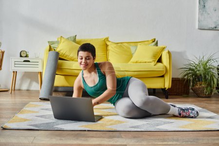 joyous woman with short hair lying on floor and looking at laptop during remote sport lesson