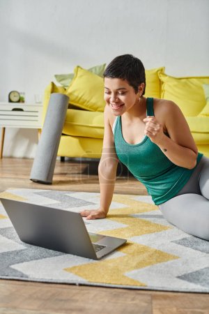 vertical shot of jolly woman with tattoo smiling at laptop screen with remote sport lesson, fitness