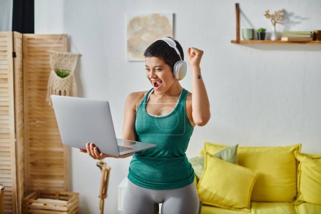young pretty woman with headphones cheering and smiling at laptop camera at remote fitness class