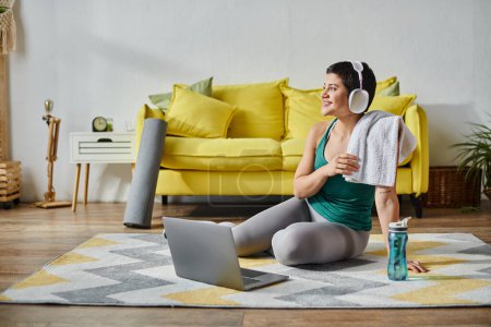 sportliche Frau auf dem Boden sitzend mit Handtuch und Kopfhörer neben Laptop und Wasserflasche, Fitness