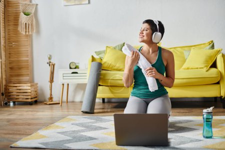 cheerful woman in sportswear sitting on floor near water bottle and laptop at remote fitness class