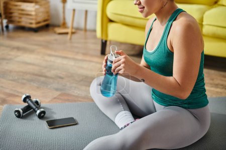 cropped view of young woman sitting on floor near dumbbells and phone with water bottle in hands