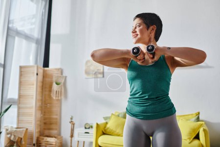 cheerful attractive woman with short hair exercising with dumbbells and looking away, fitness
