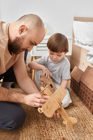 Foto de Tiro vertical de barbudo padre moderno pasar tiempo con su hijo pequeño jugando juguetes de madera - Imagen libre de derechos
