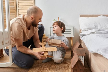 Foto de Alegre padre moderno e hijo en acogedora ropa de casa jugando con juguetes de madera y mirándose el uno al otro - Imagen libre de derechos