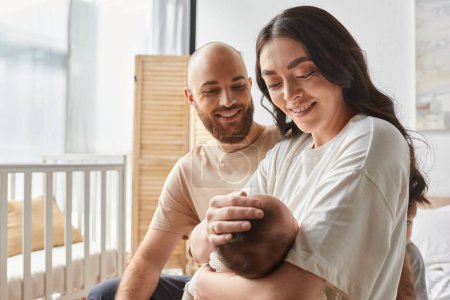 cheerful modern parents in cozy homewear sitting together on bed holding newborn baby, family