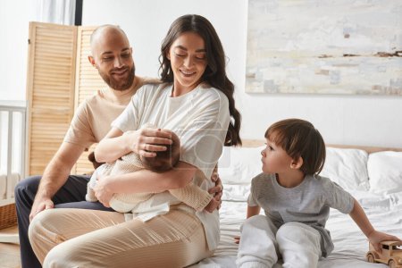 joyous parents sitting on bed with newborn baby in arms with their little son sitting next to them
