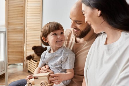 joyous little boy with wooden toy car in hands smiling at his loving parents sitting on laps, family