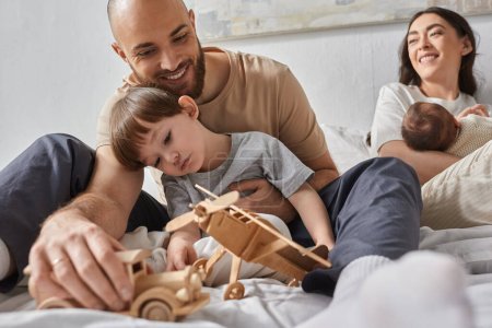 freudiger Vater, der mit seinem kleinen Sohn mit Spielzeug spielt, während seine Frau sie mit dem kleinen Jungen ansieht