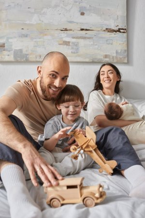 Fröhlicher bärtiger Vater umarmt seinen Sohn lächelnd in die Kamera mit seiner Frau und dem neugeborenen Baby vor der Kulisse