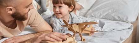alegre padre acostado en la cama con su pequeño hijo jugando juguetes de madera y sonriendo el uno al otro, pancarta