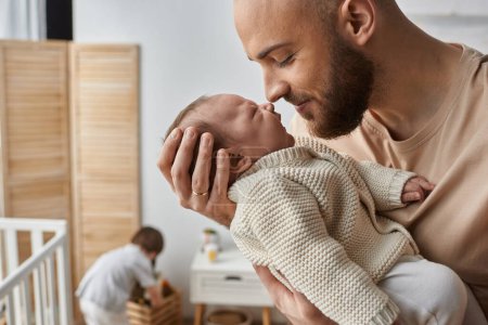 Photo for Focus on father holding lovingly his newborn baby with his little son playing on backdrop, family - Royalty Free Image