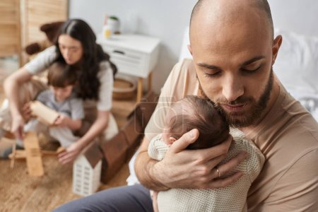 Photo for Loving father hugging his newborn baby with blurred mother and little son on backdrop, family - Royalty Free Image