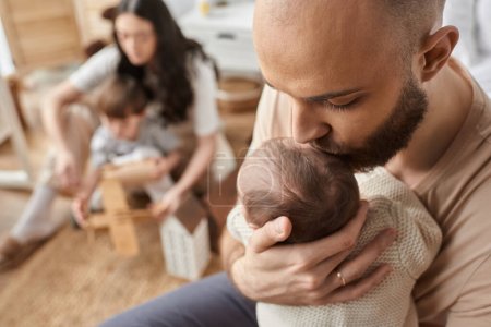 Photo for Focus on bearded father kissing his baby in forehead with his blurred wife and son on backdrop - Royalty Free Image