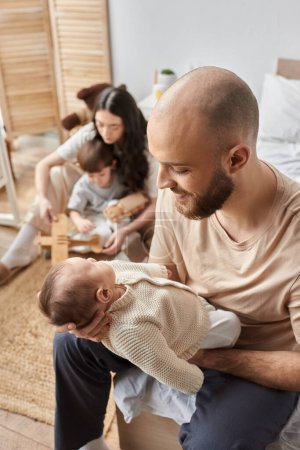 centrarse en el padre mirando con amor a su bebé recién nacido con la esposa borrosa y el pequeño hijo en el telón de fondo